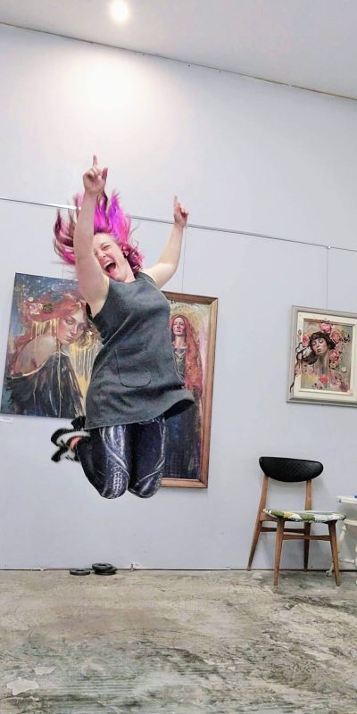 Sara Léger in Cherry Bomb Studio, captured mid-jump in front of her expressive, figurative artwork.